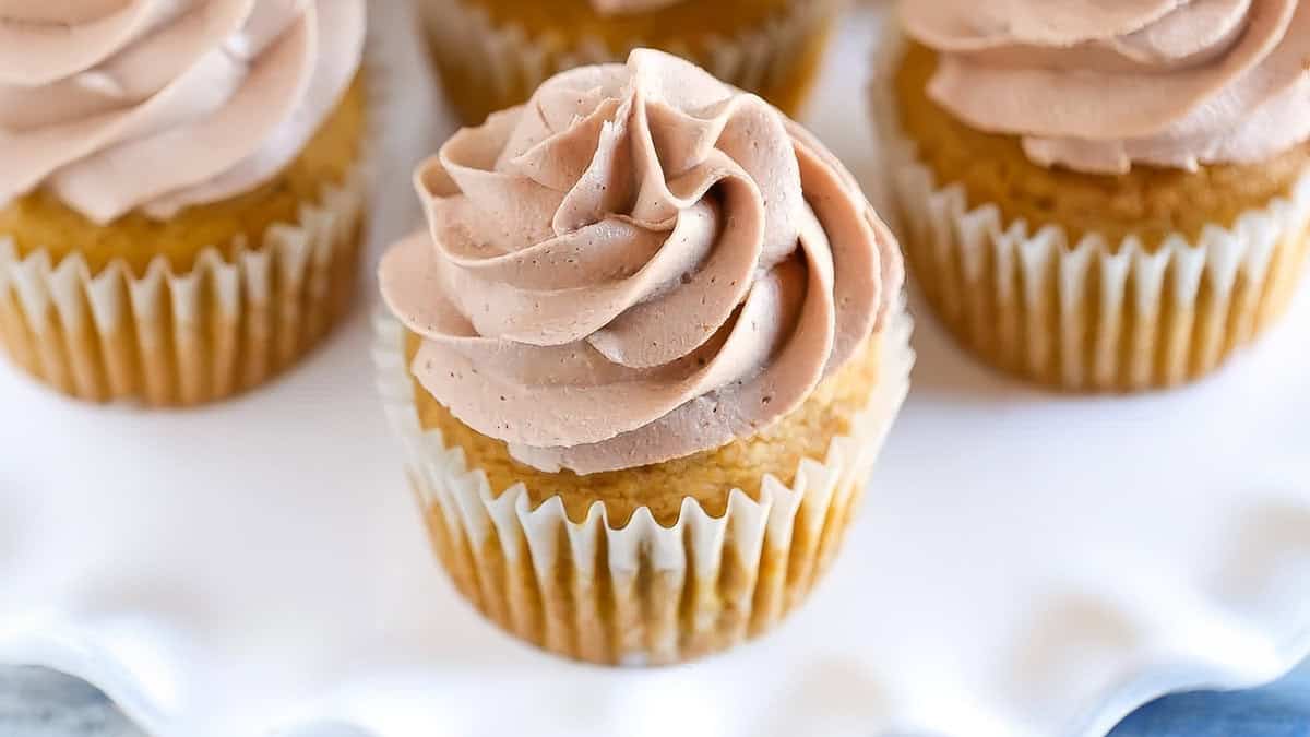 A close-up of desserts featuring vanilla cupcakes with swirled chocolate frosting on top, placed on a white surface. The baked goods are in white paper liners, with the frosting arranged in a tall, decorative spiral.