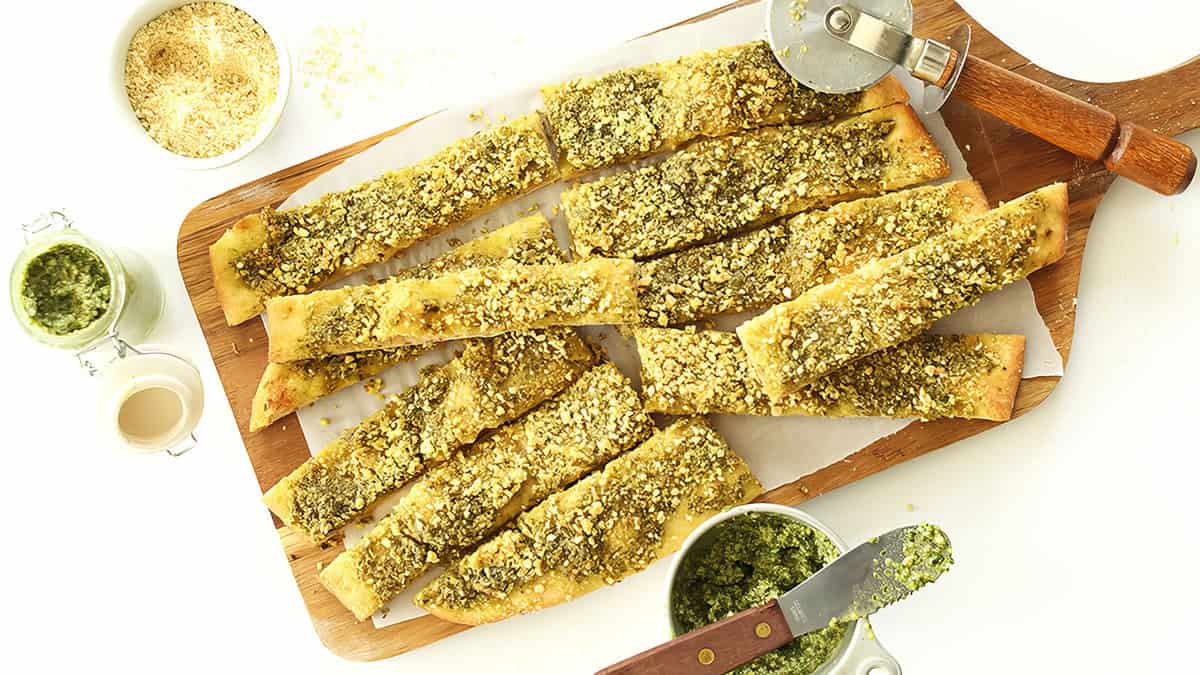 A wooden board topped with several slices of golden-brown pesto garlic breadsticks, surrounded by small bowls of grated cheese, pesto sauce, and a pizza cutter. Some breadsticks have a vibrant green pesto spread and are sprinkled with grated cheese—an irresistible addition to your favorite pesto recipes.