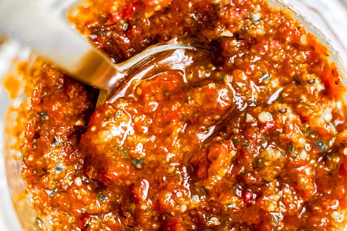 A close-up of a spoon scooping chunky homemade chili paste. The vibrant mixture includes visible pieces of red chilies, seeds, and spices, glistening with a slight oily sheen. The texture appears thick and coarse, suggesting a rich and flavorful condiment perfect for enhancing your recipes.