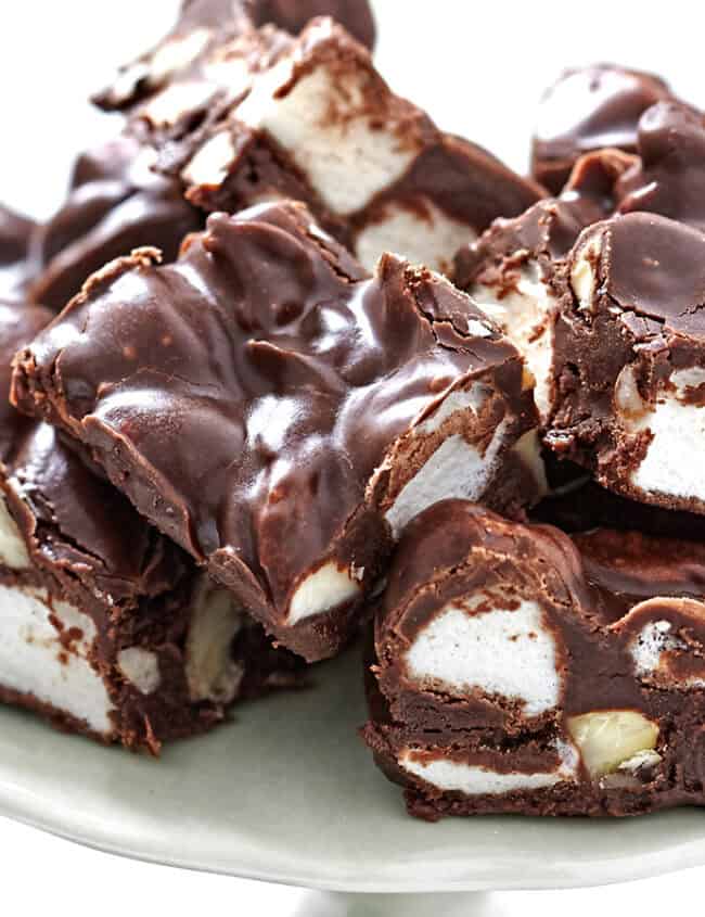 A plate of rocky road fudge is piled high with chunks of chocolate, marshmallows, and nuts. The fudge has a glossy, rich brown appearance and is densely packed with large pieces of marshmallows and nuts. A must-try from delicious recipes, the plate is set against a white background.