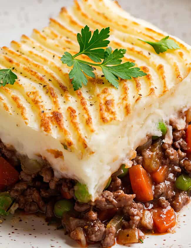 A plate of shepherd's pie topped with a layer of golden-brown mashed potatoes, garnished with parsley. This easy ground beef recipe includes a filling of ground meat, diced carrots, peas, and a rich brown sauce, all served on a white speckled plate.