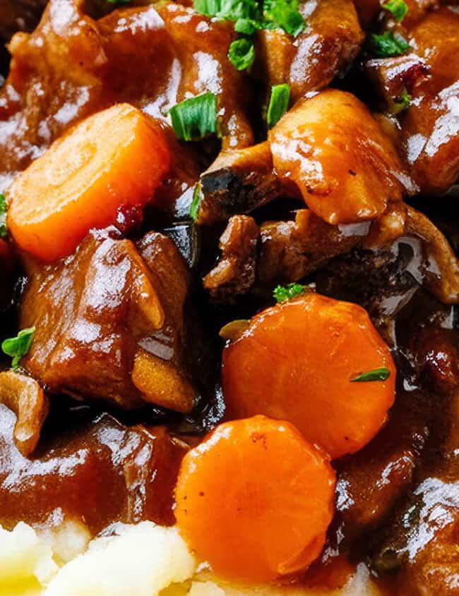 Close-up of a savory beef stew with chunks of beef, carrots, and vegetables in a thick brown gravy, served over creamy mashed potatoes. This indulgent entry from our Beef Recipes collection is garnished with chopped green herbs.