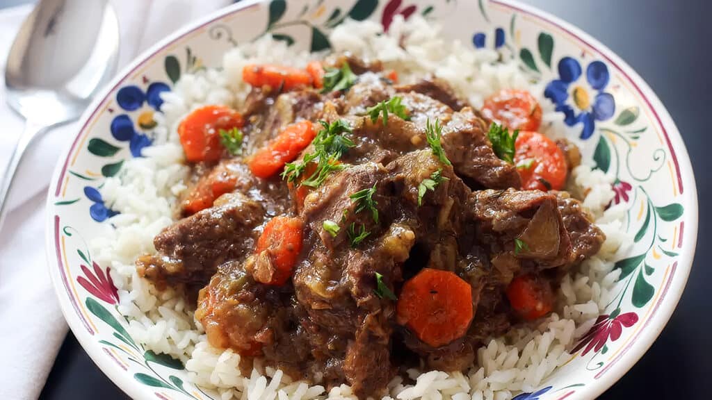 A colorful bowl filled with white rice topped with a savory beef stew containing tender beef chunks, sliced carrots, and garnished with fresh parsley. The rich and flavorful stew reflects the beauty of Slow Cooker Recipes, set against a floral-patterned bowl, with a spoon on the side.