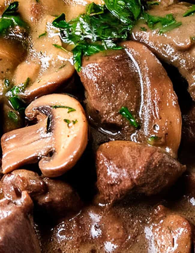 Close-up of a hearty mushroom and beef stew, perfect for slow cooker recipes. The dish features chunks of beef and sliced mushrooms in a rich, dark gravy, garnished with fresh chopped parsley. A wooden spoon is partially submerged in the stew, suggesting serving or stirring.