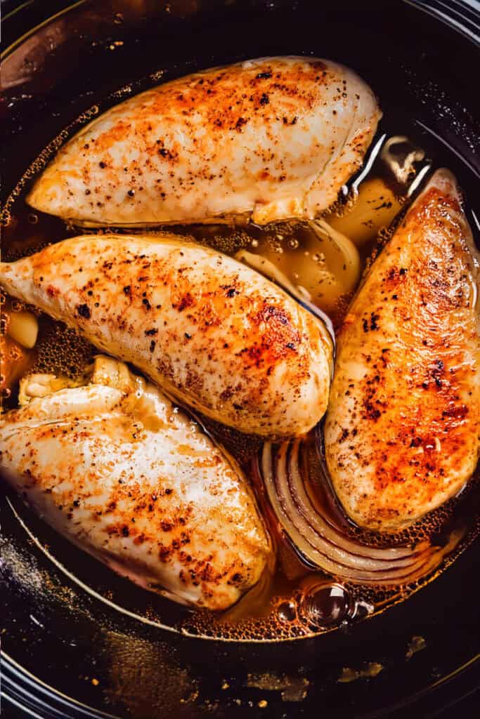 Four seasoned chicken breasts cook in a dark-colored pan with a savory broth. The chicken is browned and surrounded by thin slices of onion, evoking the rich flavors of a slow-cooker dish.