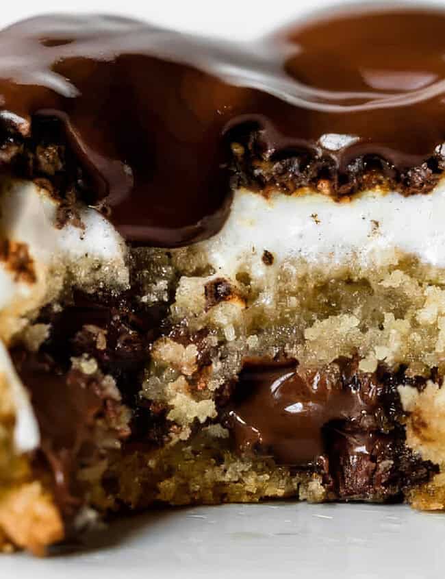 A close-up shot of a cookie with a bite taken out of it reveals layers of gooey chocolate, sticky marshmallows, and a crumbly base. The rich chocolate topping glistens, adding a glossy finish to this sticky and sweet treat from the realm of delicious recipes.