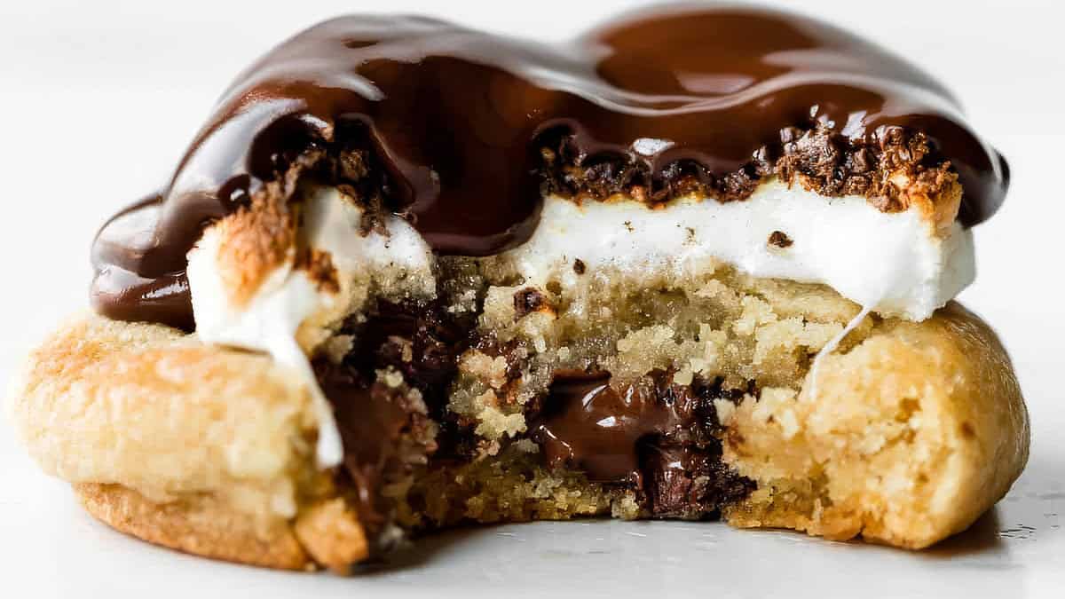 A close-up shot of a cookie with a bite taken out of it reveals layers of gooey chocolate, sticky marshmallows, and a crumbly base. The rich chocolate topping glistens, adding a glossy finish to this sticky and sweet treat from the realm of delicious recipes.