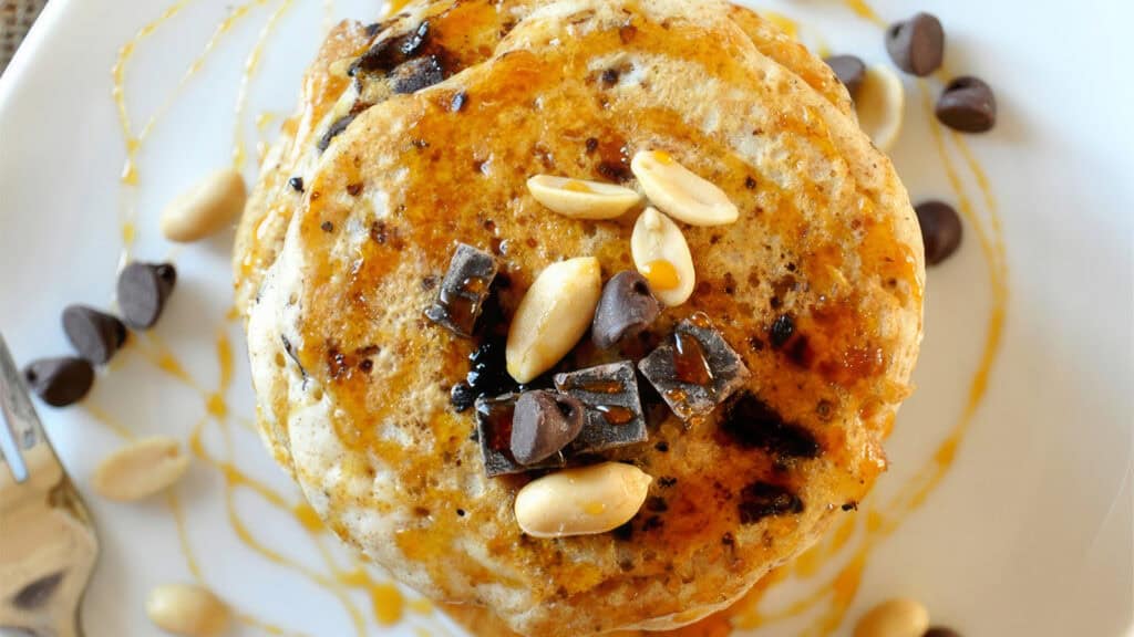 A close-up view of two pancakes on a white plate, drizzled with syrup and topped with chocolate pieces and peanuts. Chocolate chips and peanuts are scattered around on the plate, providing inspiration for your next pancake recipes. The edges of a fork are partially visible on the left side.