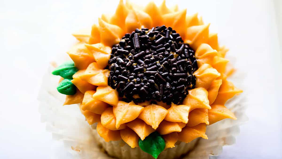 A delightful cupcake decorated to resemble a sunflower, with vibrant yellow petals made of icing. The center is filled with chocolate sprinkles, and there are small green leaves made of icing on the sides. This charming baked good sits in a white paper liner, perfect for any desserts lover.