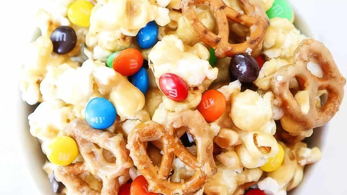 A close-up view of a snack mix in a bowl, consisting of popcorn, pretzels, colorful candy-coated chocolates, and marshmallows. The sticky and sweet mix appears coated with a light glaze, giving it a shiny appearance.