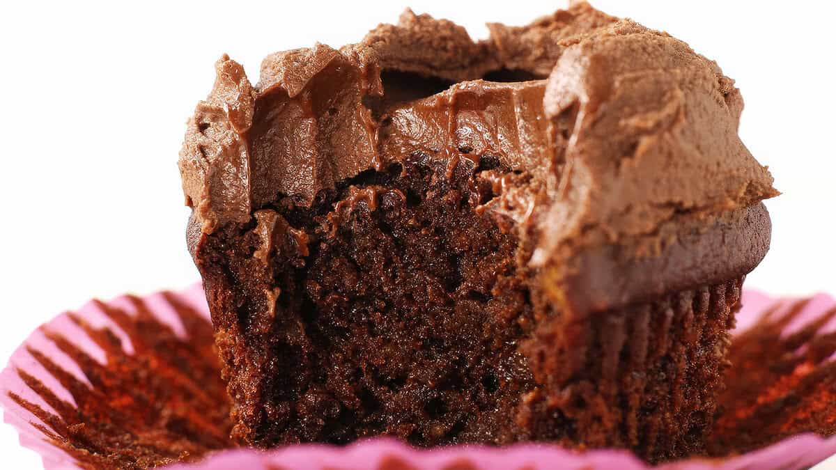 A chocolate cupcake with rich, creamy chocolate frosting, partially eaten, sits on an opened pink cupcake liner. The texture of the moist cake and fluffy frosting epitomizes delightful dessert baked goods.