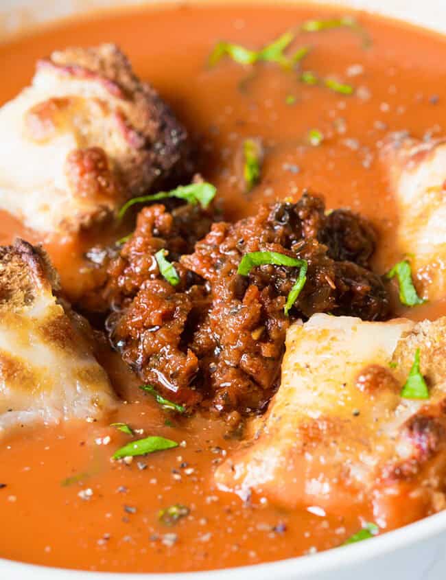 Close-up of a bowl of tomato soup garnished with browned croutons and a dollop of ground meat mixture in the center. Fresh chopped herbs and a hint of pesto are sprinkled on top, adding color and freshness. The creamy soup contrasts with the toasted texture of the croutons, making it perfect cooking inspiration for new recipes.