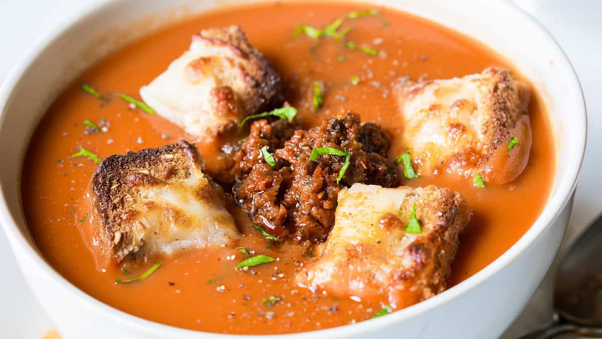 Close-up of a bowl of tomato soup garnished with browned croutons and a dollop of ground meat mixture in the center. Fresh chopped herbs and a hint of pesto are sprinkled on top, adding color and freshness. The creamy soup contrasts with the toasted texture of the croutons, making it perfect cooking inspiration for new recipes.