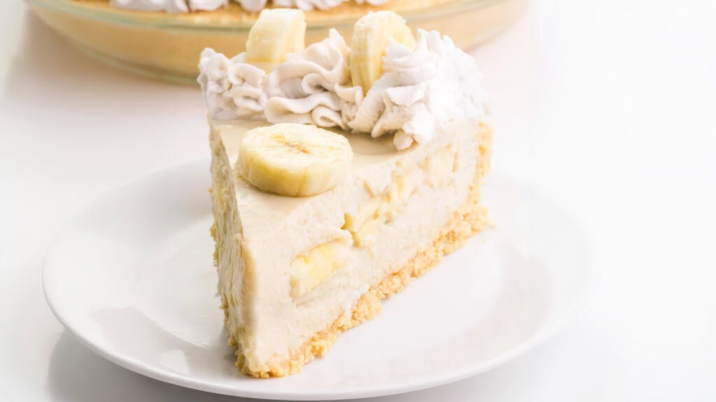 A slice of banana cream pie on a white plate. The pie features a creamy filling with banana slices and a crumbly crust. The top is garnished with whipped cream and additional banana slices. Perfect for fans of banana recipes, the image highlights a larger pie in the background.