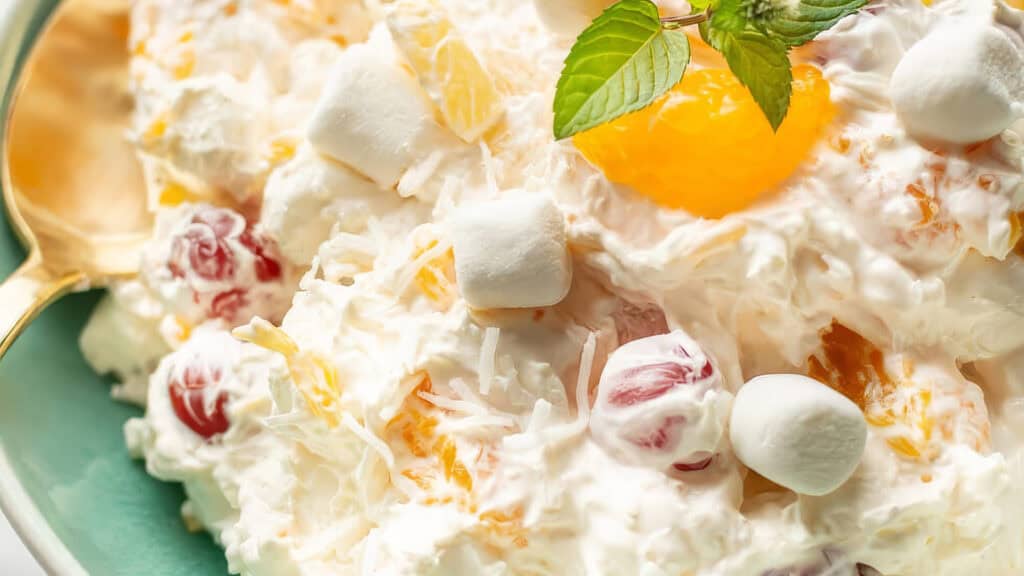 A close-up view of a creamy ambrosia salad, showcasing a mixture of marshmallows, mandarin orange slices, pineapple chunks, red maraschino cherries, and grated coconut, all garnished with a fresh mint sprig. A gold-colored spoon is partially visible on the left side—a true staple in Southern recipes.