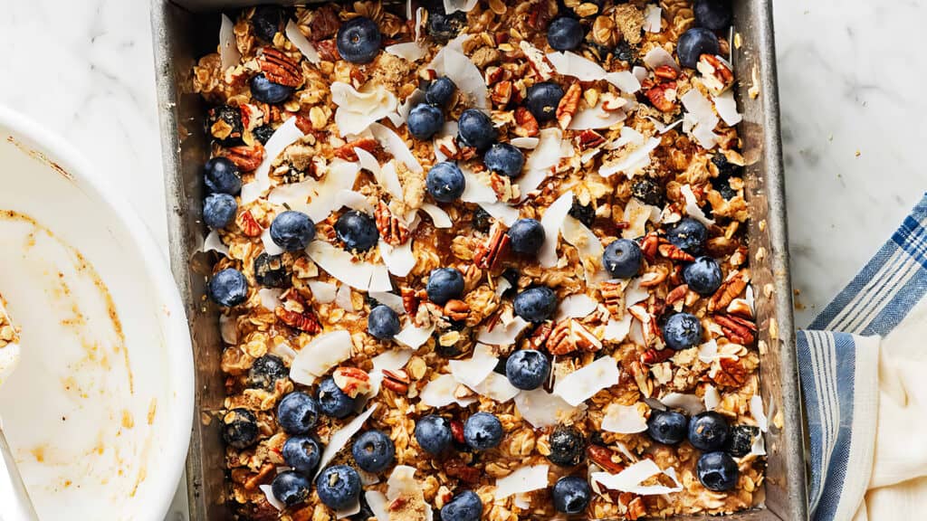 A square baking dish filled with baked oatmeal topped with blueberries, coconut flakes, and pecans is placed on a marble surface. A blue-striped white cloth is visible on the right side of the dish, making it a delightful addition to your favorite blueberry recipes.