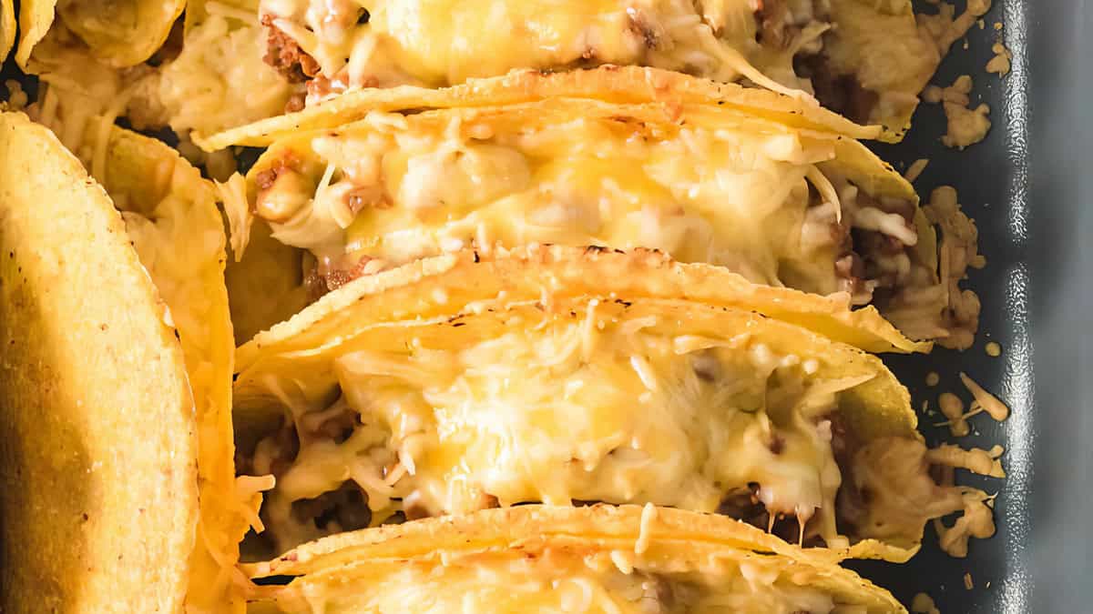 A close-up view of a baking dish filled with crunchy tacos. The tacos are generously stuffed with beef and topped with melted cheese, creating a gooey and appetizing appearance. 