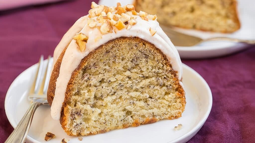 A slice of banana nut cake with white icing and chopped nuts on top, served on a white plate with a fork. Another slice is visible in the background. The tablecloth is purple, adding a touch of color to the setting—a delightful treat for those who love Bundt cake recipes.