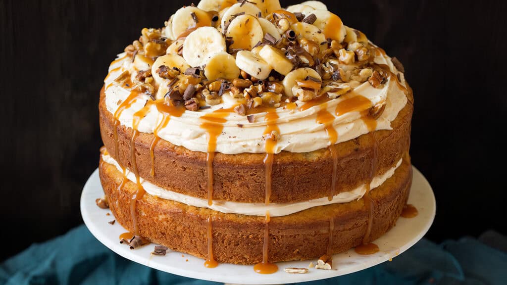 A layered cake topped with creamy frosting, banana slices, chopped nuts, and caramel drizzles. The cake has two thick layers with a generous amount of frosting between them, inspired by the best caramel recipes. It is displayed on a white cake stand against a dark backdrop.