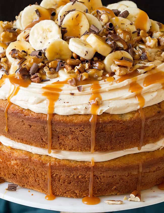 A layered cake topped with creamy frosting, banana slices, chopped nuts, and caramel drizzles. The cake has two thick layers with a generous amount of frosting between them, inspired by the best caramel recipes. It is displayed on a white cake stand against a dark backdrop.