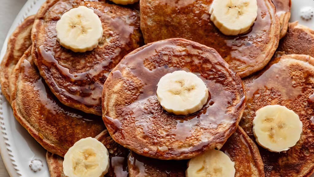 A close-up of a stack of golden-brown pancakes topped with banana slices and drizzled with syrup. The fluffy, moist pancakes create an appetizing breakfast scene, perfect for trying out new pancake recipes. The banana slices add a fresh touch to the dish.