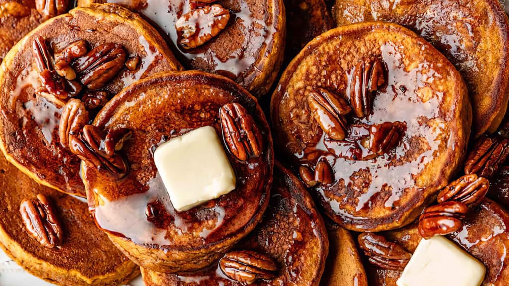 A close-up of a stack of fluffy pancakes topped with melting butter, glistening maple syrup, and pecans. The golden brown pancakes and sumptuous toppings create a mouth-watering appearance, perfect for any pancake recipe lover.