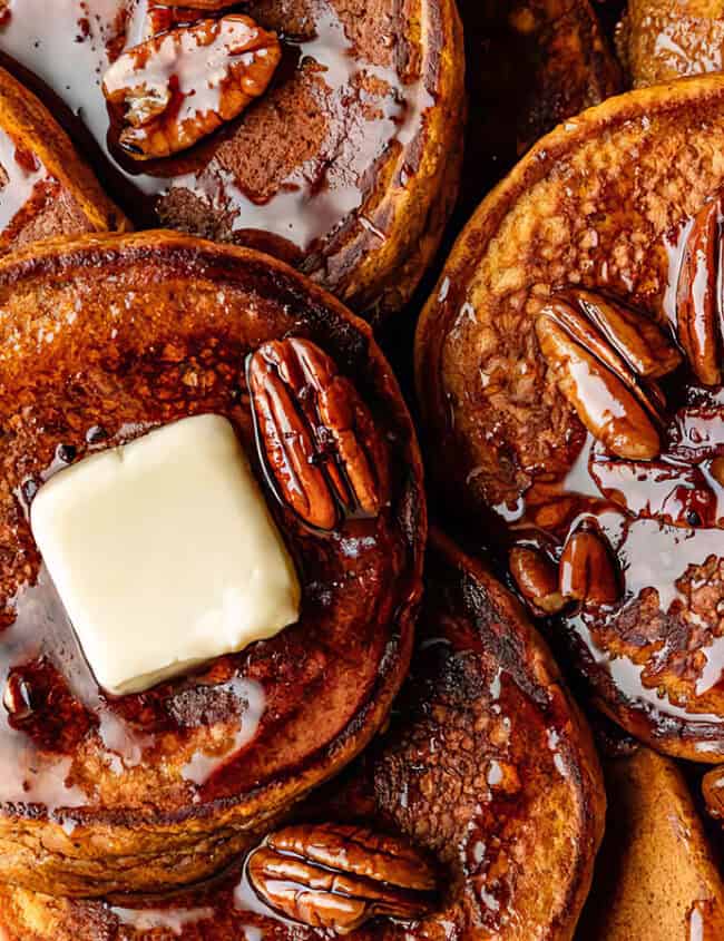 A close-up of a stack of fluffy pancakes topped with melting butter, glistening maple syrup, and pecans. The golden brown pancakes and sumptuous toppings create a mouth-watering appearance, perfect for any pancake recipe lover.