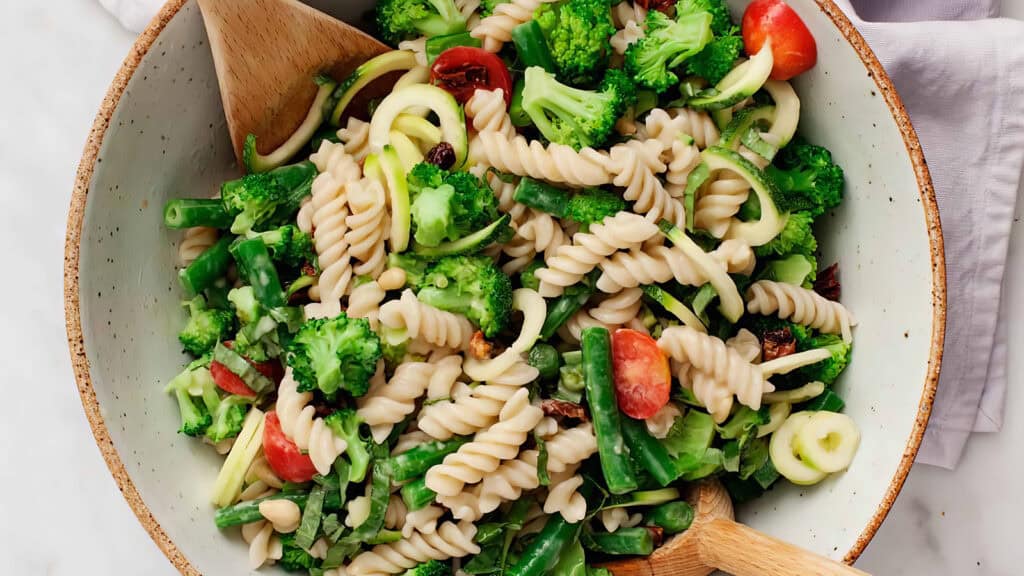 A bowl of colorful pasta salad featuring rotini noodles, broccoli florets, cherry tomato halves, green beans, zucchini slices, and mixed greens. Wooden utensils are placed in the bowl, ready to serve. This vibrant and fresh dish is a highlight among pasta salad recipes, sitting on a light background.