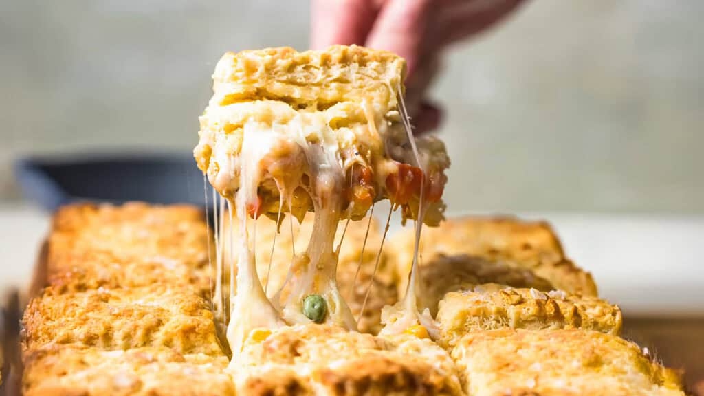 A hand is lifting a piece of Cheesy Chicken Pot Pie with Biscuit Topping from a baking pan. 