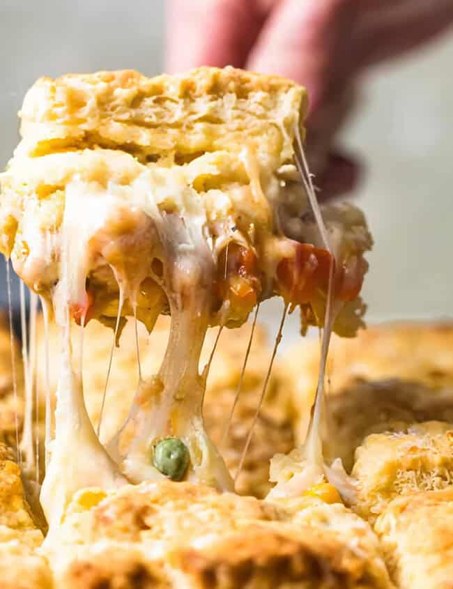 A hand is lifting a piece of Cheesy Chicken Pot Pie with Biscuit Topping from a baking pan.