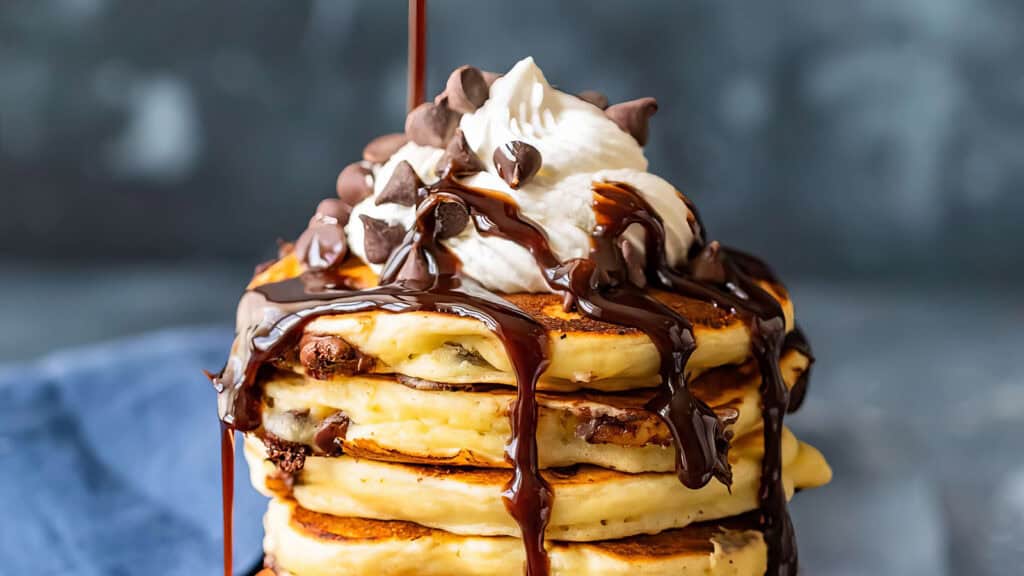 A stack of fluffy pancakes topped with a generous dollop of whipped cream, chocolate chips, and drizzled with rich chocolate syrup—a perfect addition to your favorite chocolate chip recipes. The background is out of focus, making the pancakes the central focus of the image.