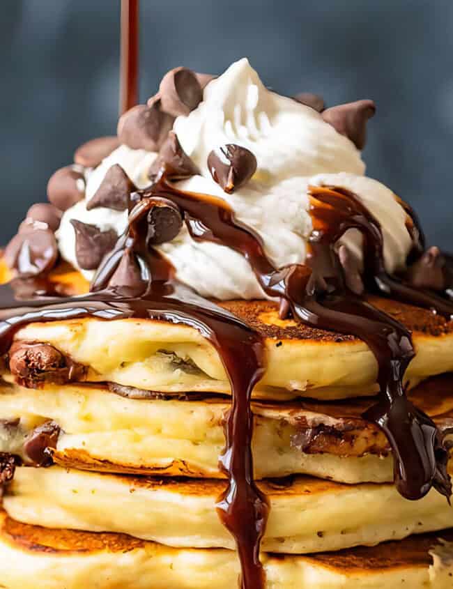 A stack of fluffy pancakes topped with a generous dollop of whipped cream, chocolate chips, and drizzled with rich chocolate syrup—a perfect addition to your favorite chocolate chip recipes. The background is out of focus, making the pancakes the central focus of the image.