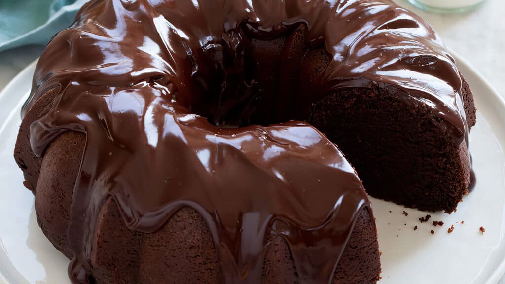 A rich chocolate bundt cake is shown on a white plate. The cake, a star among bundt cake recipes, is topped with glossy chocolate glaze that drips down the sides. A single slice has been cut from the cake, revealing its moist and dense interior.