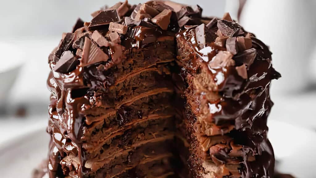 A decadent multi-layer chocolate cake with rich, dark chocolate frosting and large chocolate chunks on top is shown. Several layers are visible with a slice cut out, revealing the moist and dense interior. Perfect for indulgent chocolate chip recipes, the background is blurred to focus attention on the cake.