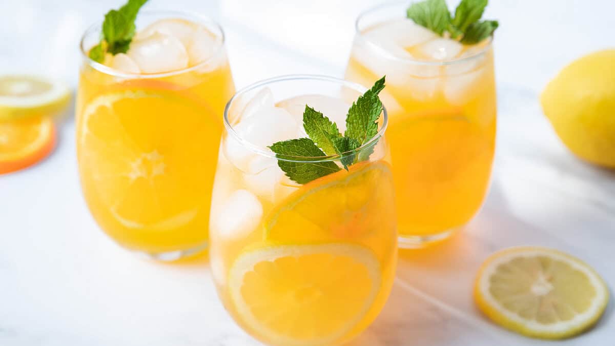 Three glasses of iced lemonade garnished with mint leaves and lemon slices are displayed on a white surface. In the background, a whole lemon and lemon slices can be seen. The refreshing drinks are presented in a bright, inviting manner, reminiscent of classic cocktail recipes.