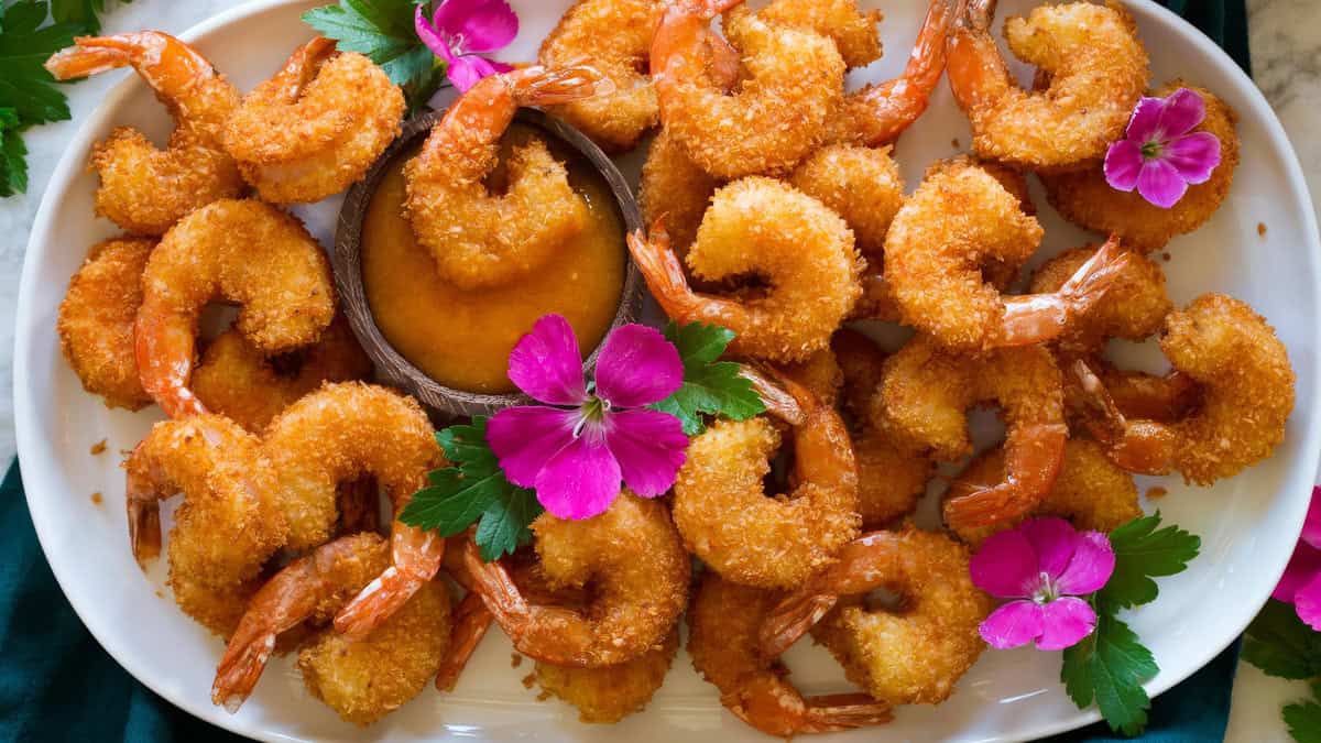 A white plate filled with breaded fried shrimp recipes arranged around a small dish of orange dipping sauce in the center. The shrimp are garnished with green herbs and vibrant pink flowers, creating a colorful and appetizing presentation.