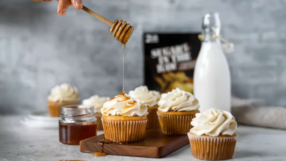 A hand drizzles honey onto a cupcake topped with creamy frosting using a wooden honey dipper. Nearby, more frosted cupcakes are arranged on a wooden board, highlighting the delightful array of desserts. A jar of honey and a bottle of milk are in the background, creating a cozy, dessert-themed setting.