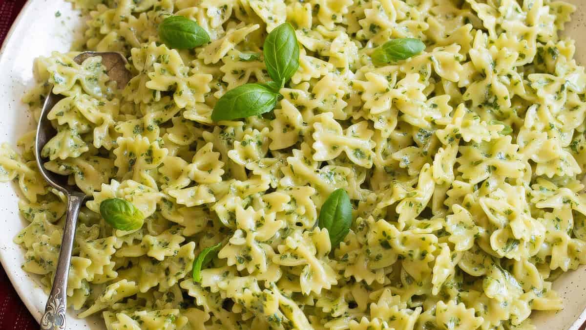 A plate of bow-tie pasta is coated in a creamy pesto sauce, garnished with fresh basil leaves. A metal serving spoon rests in the pasta, ready to serve. The dish is colorful and appetizing with a bright green hue from the pesto—perfect for those exploring new pesto recipes.