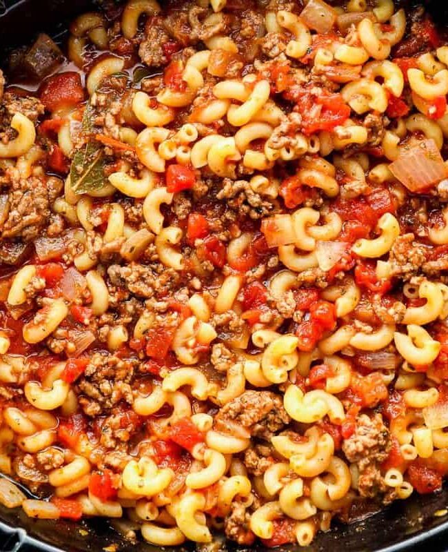 A close-up of a slow cooker filled with a hearty dish of cooked macaroni pasta mixed with ground beef, tomatoes, onions, and spices. The ingredients are well combined, creating a colorful and savory meal that's perfect for fans of ground beef recipes.