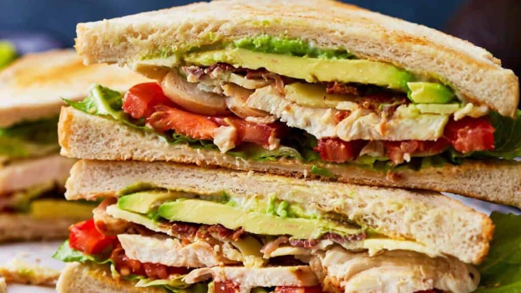 A close-up of a stacked sandwich cut in half, showcasing layers of toasted bread, chicken slices, avocado, lettuce, tomato, and crispy bacon. Perfect for picnic recipes, the sandwich appears fresh and appetizing with vibrant colors of the vegetables and well-cooked ingredients.