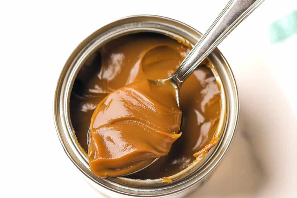 An open can of dulce de leche with a spoonful of the caramel-colored, creamy spread being lifted out. The surface of the dulce de leche in the can is smooth and glossy, reminiscent of a luscious salted caramel treat. The background is white and slightly blurred.