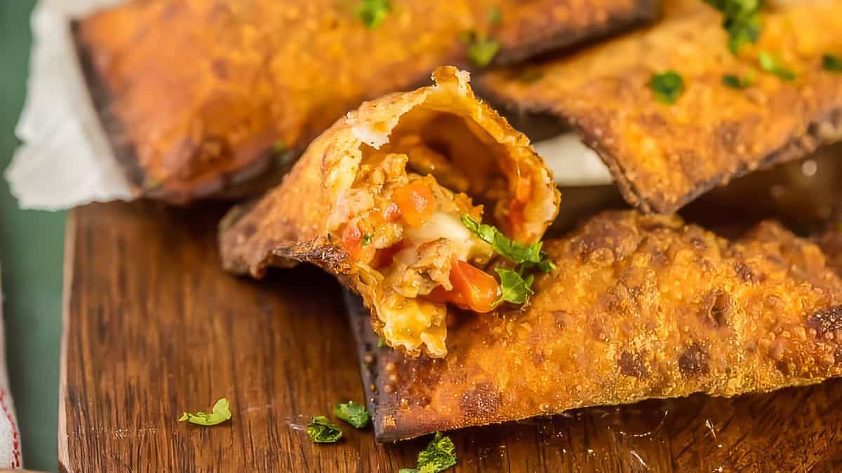 Close-up of a crispy egg roll split open to reveal a filling of seasoned ground beef, diced red bell peppers, and melted cheese. The egg roll is sprinkled with chopped parsley and placed on a wooden surface with additional easy-to-make egg rolls in the background.