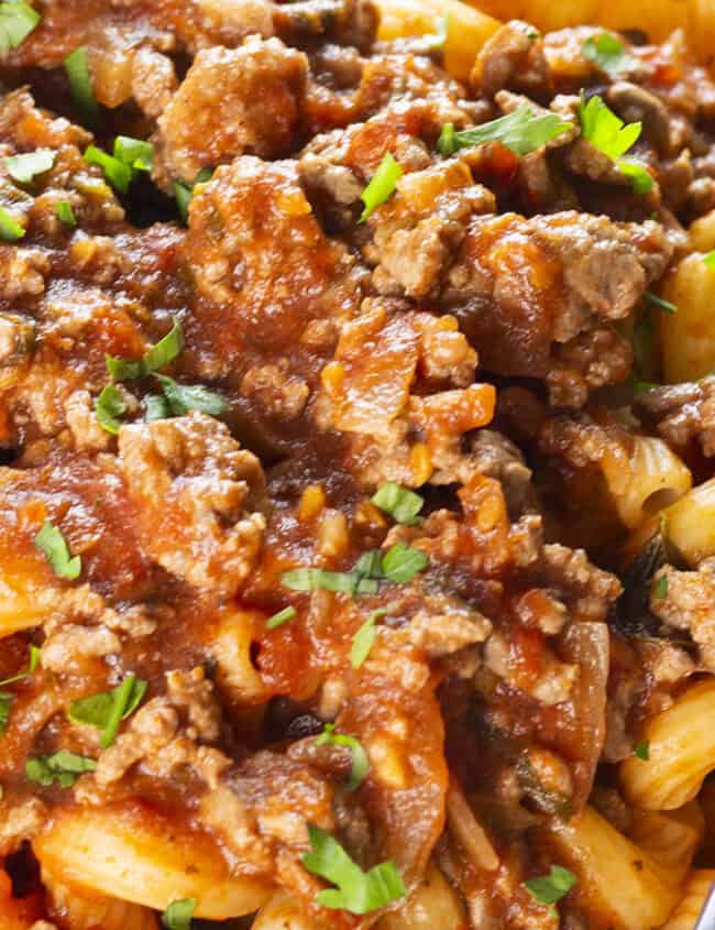 A close-up image of a plate filled with spiral pasta topped with a generous amount of rich meat sauce made from ground beef. The sauce appears hearty, with visible chunks of ground meat and sprinkled with chopped parsley. A silver fork rests on the plate's edge.