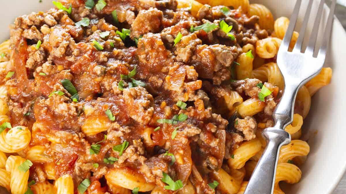 A close-up image of a plate filled with spiral pasta topped with a generous amount of rich meat sauce made from ground beef. The sauce appears hearty, with visible chunks of ground meat and sprinkled with chopped parsley. A silver fork rests on the plate's edge.