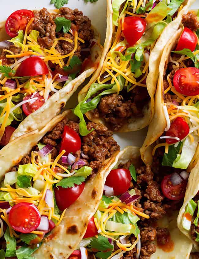 A plate of five soft shell tacos filled with seasoned ground beef, shredded cheese, chopped lettuce, diced red onions, halved cherry tomatoes, and fresh cilantro. This easy ground beef recipe sits on a gray surface with a striped cloth napkin and a small bottle of hot sauce nearby.