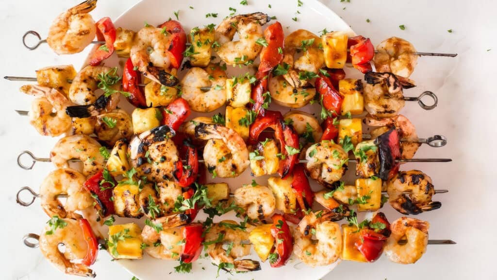 A platter of grilled shrimp kebabs with chunks of juicy pineapple, vibrant red bell peppers, and sprinkled with fresh herbs. Showcasing a colorful and appetizing presentation, this delightful creation is a gem among shrimp recipes arranged aesthetically on a white dish.