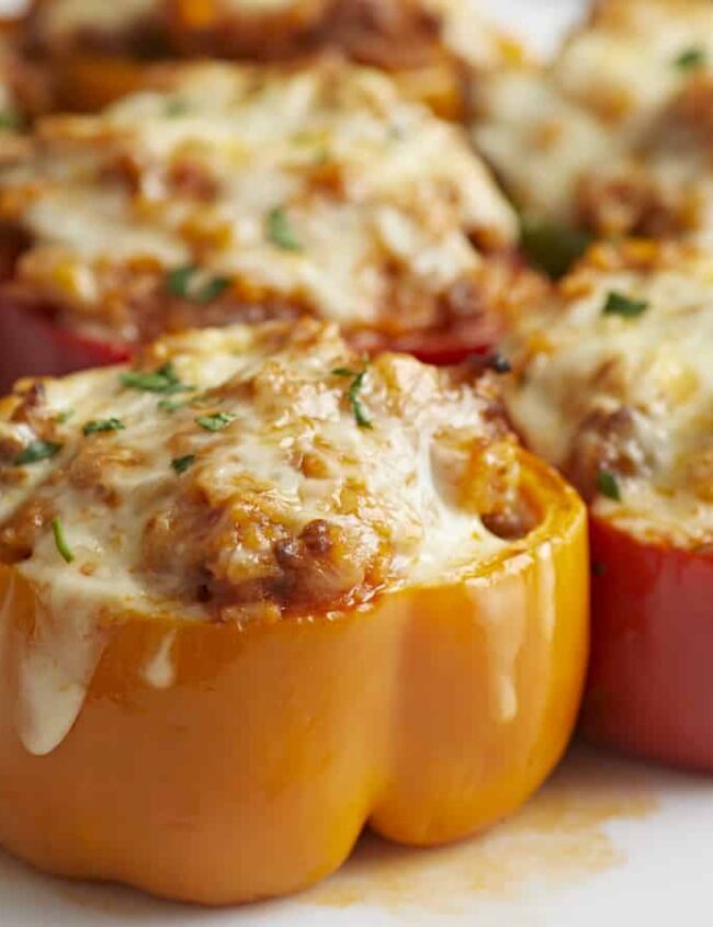 Close-up of stuffed bell peppers on a white surface. The yellow, red, and green peppers are filled with a ground meat mixture and topped with melted cheese. Some have fresh herbs sprinkled on top, with the cheese slightly browned and bubbly—a delightful dish for those exploring new peppers recipes.