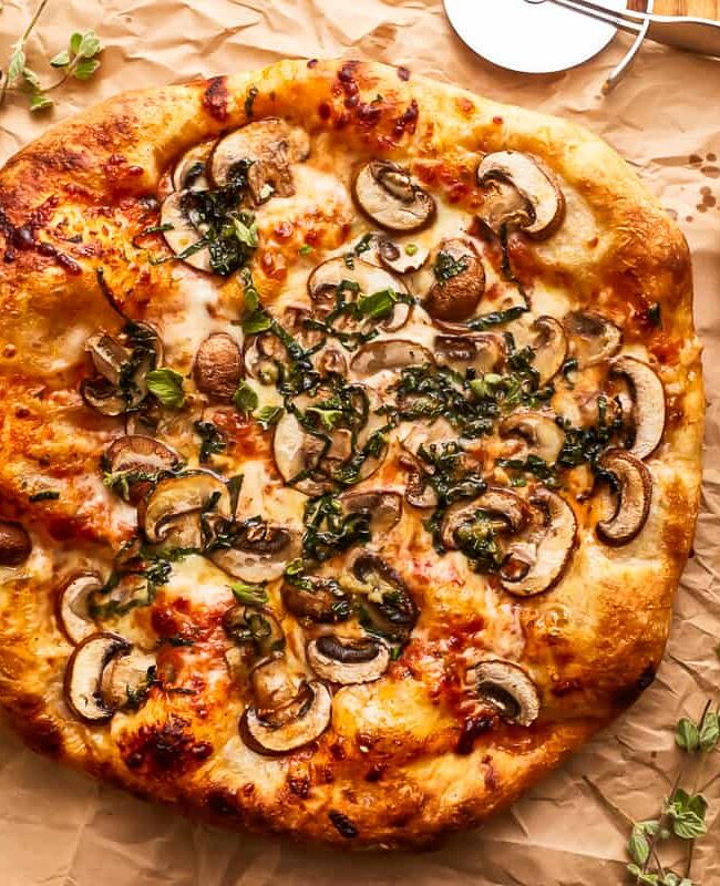 A freshly baked pizza topped with mushrooms, mozzarella cheese, and fresh herbs sits on a sheet of brown parchment paper. Perfect for mushroom recipes enthusiasts, a pizza cutter and a small bowl of olive oil with a basting brush are placed nearby on the wooden surface.