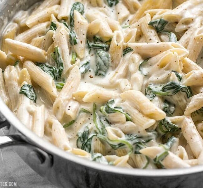 A close-up of a skillet filled with creamy spinach pasta, one of the best spinach recipes. The pasta is coated in a thick, white sauce with visible spinach leaves mixed in. The skillet is viewed from above, sitting on a light-colored countertop.