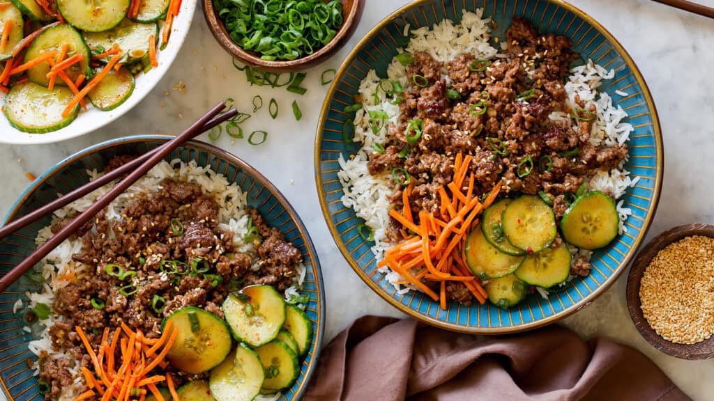 Two bowls of rice topped with seasoned ground beef, sliced cucumbers, shredded carrots, and green onions, with chopsticks resting on the side. A small bowl of sliced green onions and a bowl of sesame seeds are also visible, along with a partly visible plate of vegetables. Perfect for budget-friendly recipes!